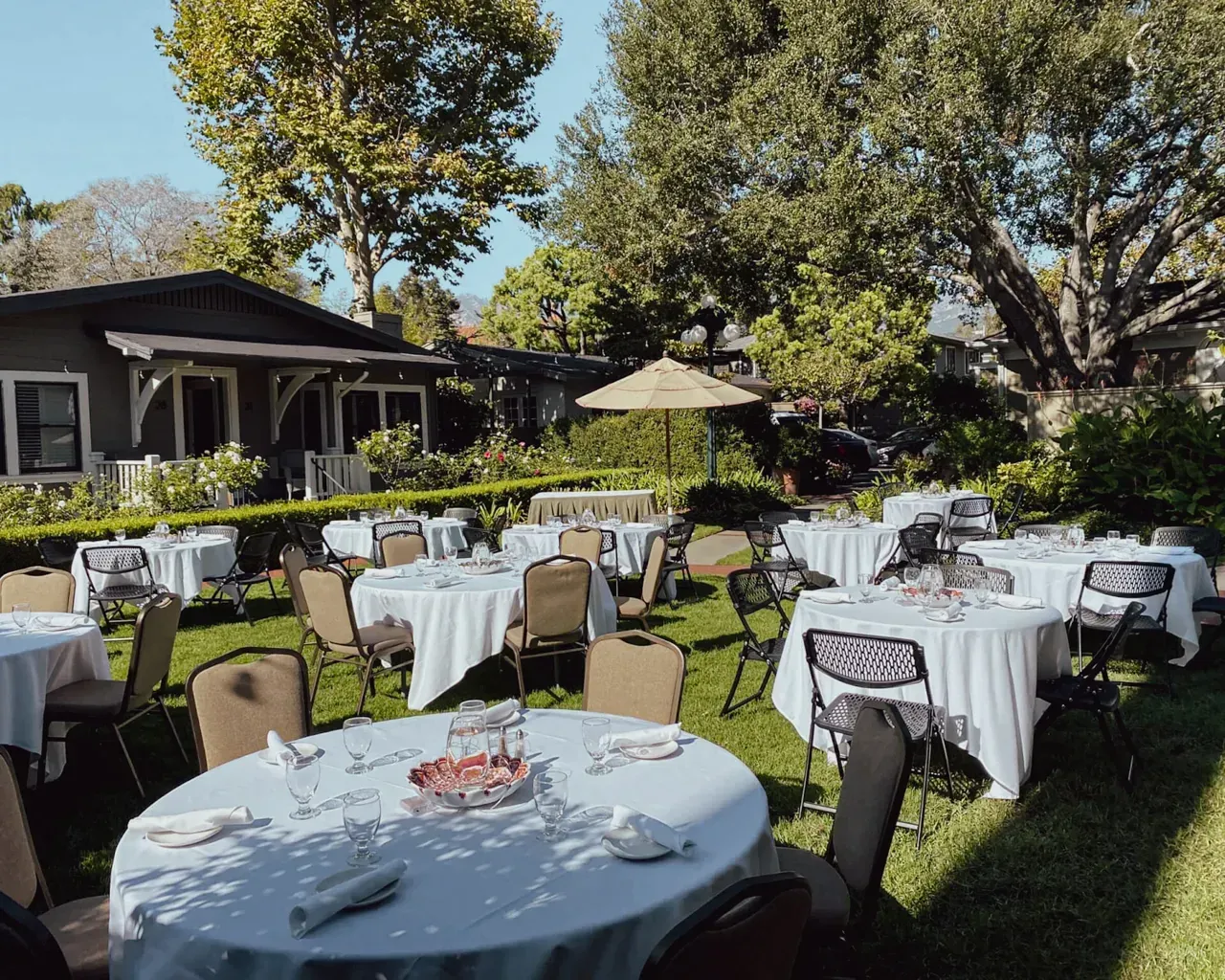 Outdoor event space with white-clothed round tables and chairs set up on a lawn in front of a house.