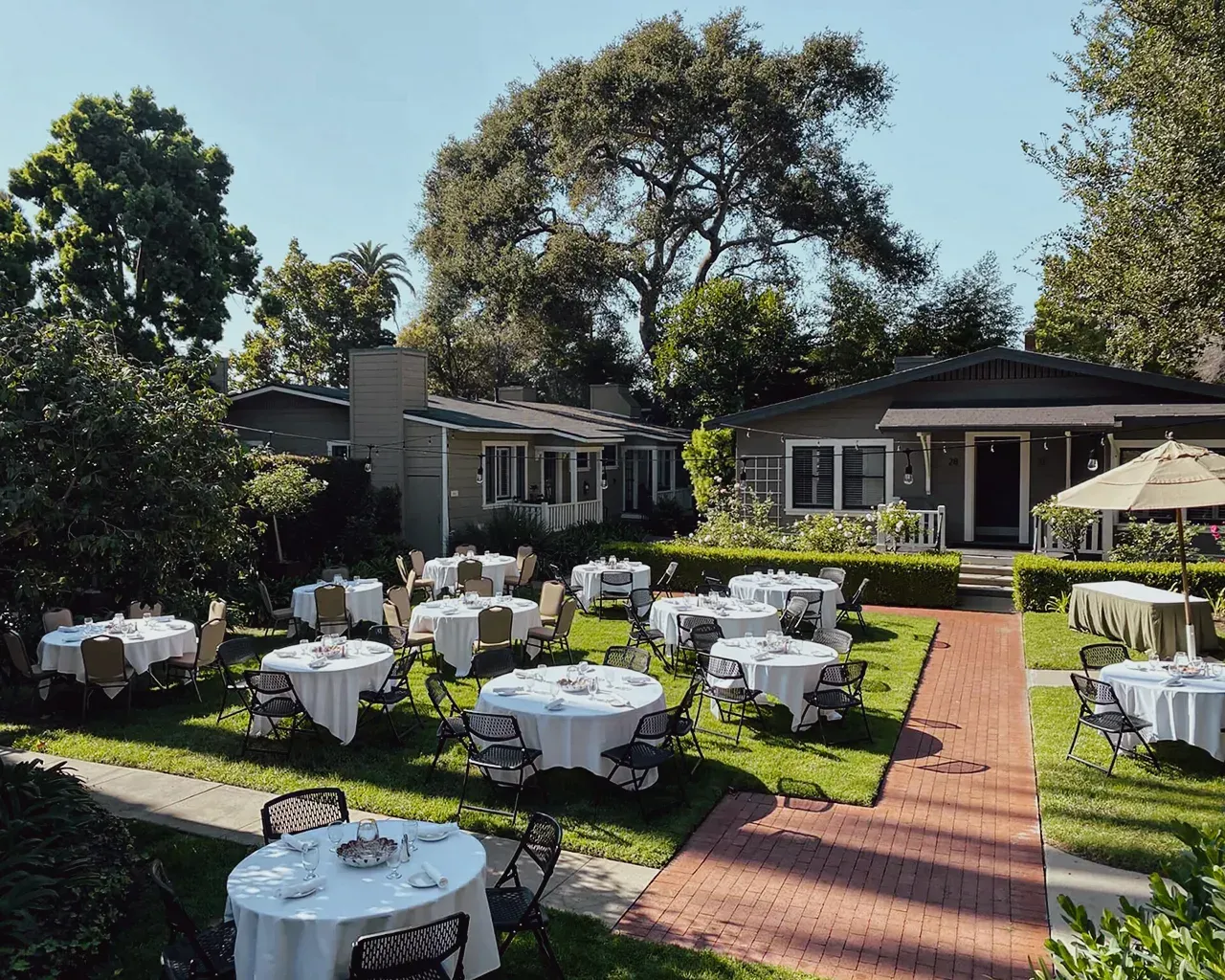 Outdoor garden setting with several round dining tables covered in white cloths, arranged on a lawn in front of homes.