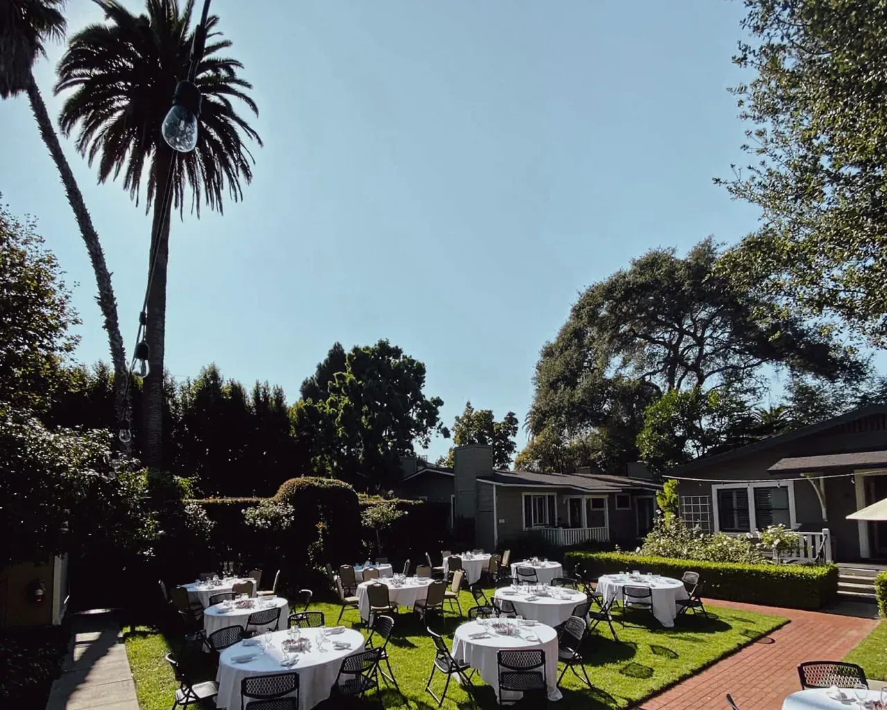 Round tables with white tablecloths and chairs set up for an outdoor garden event on a sunny day.