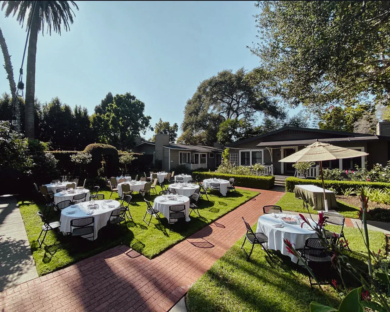 Outdoor patio area set with white-clothed round tables on a grassy lawn with a brick walkway and surrounding trees.