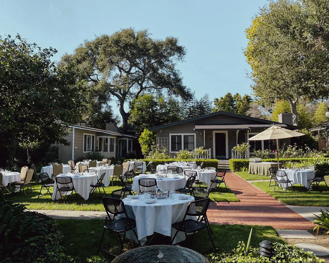 Outdoor event space with white-clothed round tables set up on a lawn in front of a house, featuring trees and a walkway.