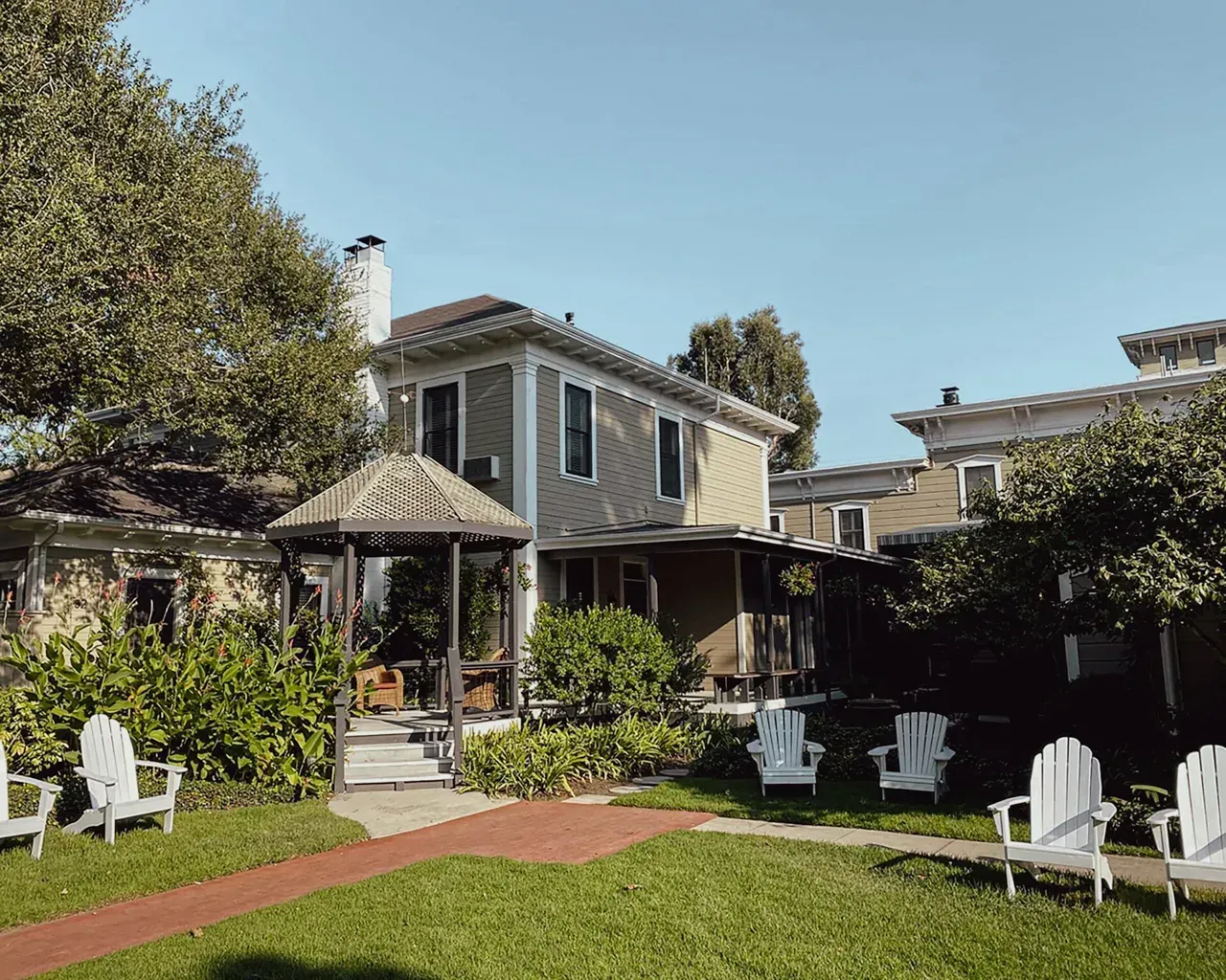 A garden with white Adirondack chairs, a gazebo, and a multi-story house under a clear blue sky.