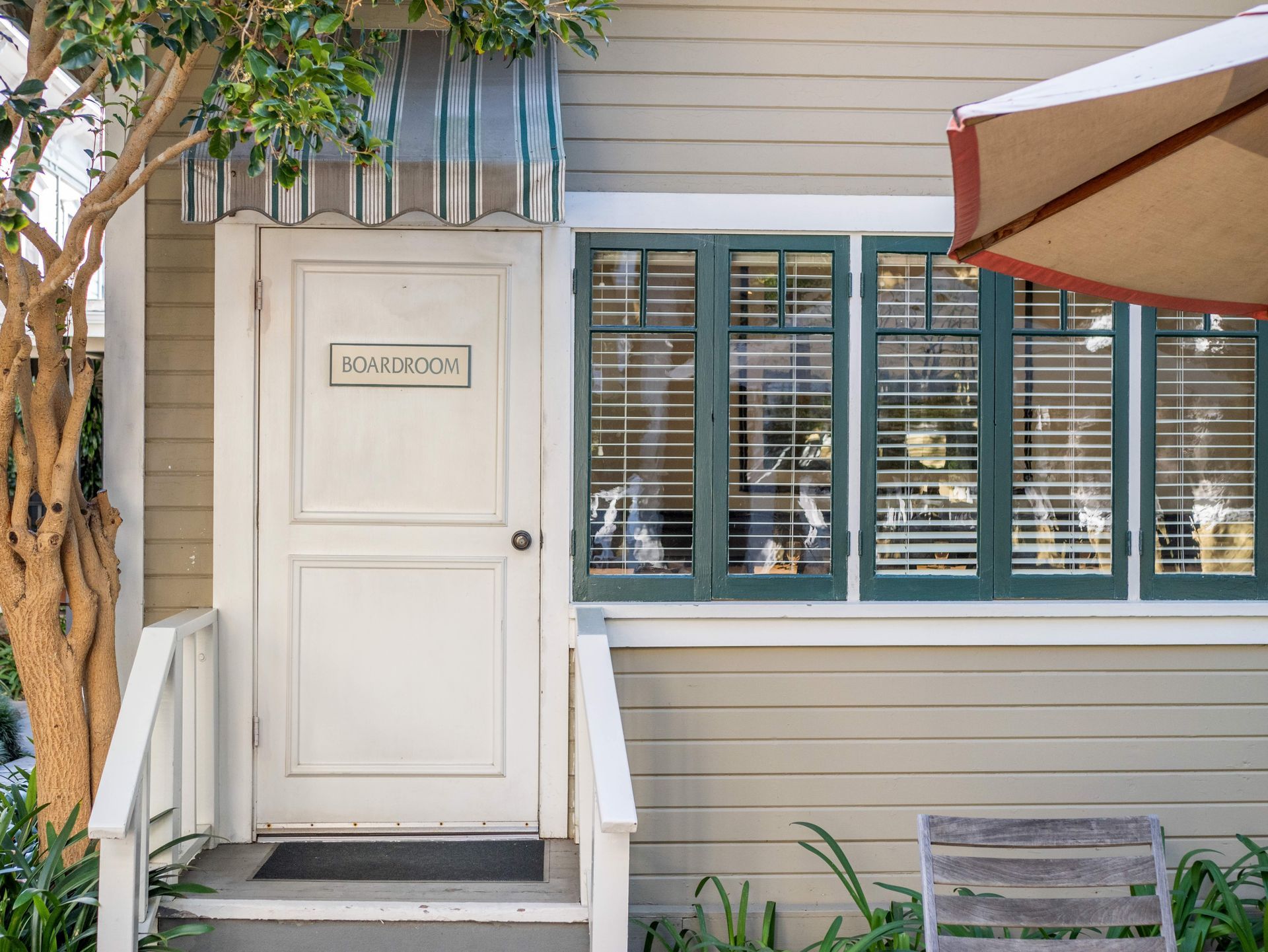 A white door with a sign and a green-framed window on a tan building, with a striped awning and a patio umbrella.