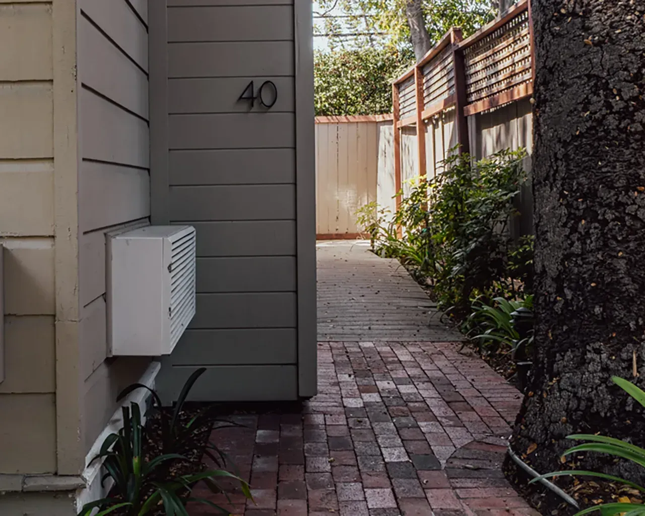 A brick walkway leads between a gray building labeled '40' and a wooden fence near a large tree.
