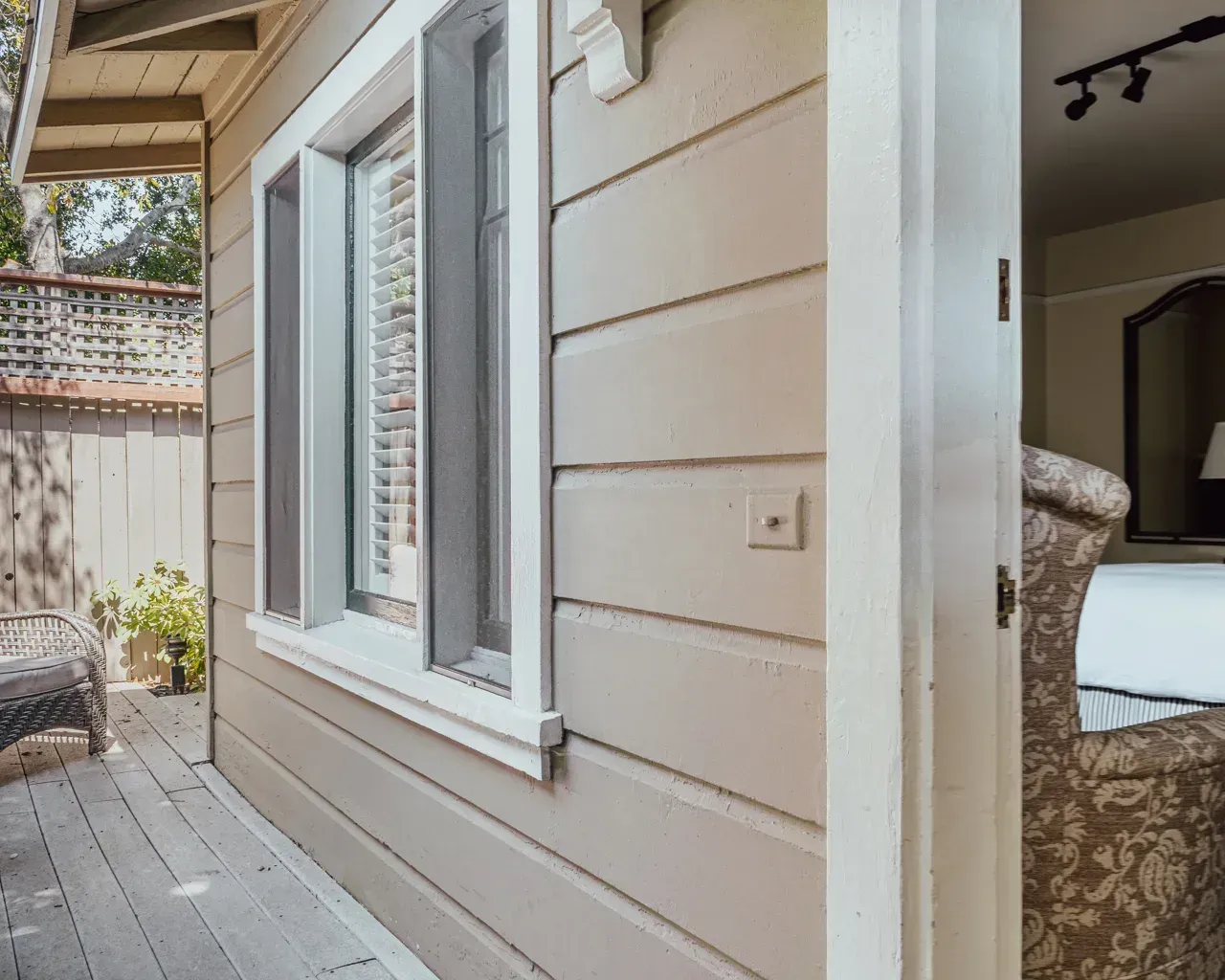 Light-tan wood siding on an exterior wall with a white-framed window next to an open doorway leading into a room.