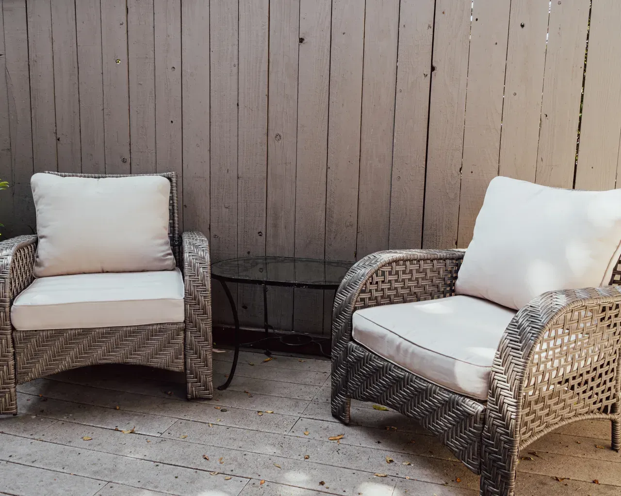 Two gray wicker armchairs with white cushions sit side-by-side on a patio in front of a wooden fence.