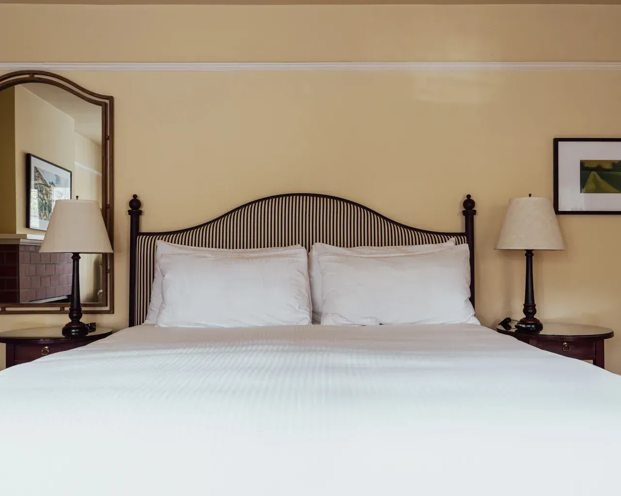 A tidy hotel bed with white linens and pillows, flanked by two wooden nightstands with lamps and framed wall art.