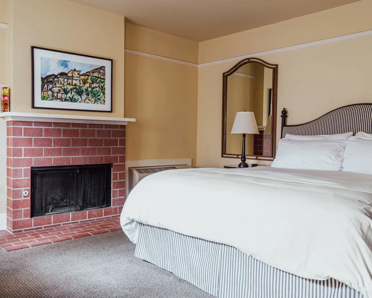 A hotel room featuring a king-size bed with striped bedding, a brick fireplace, and a mirror on a tan wall.