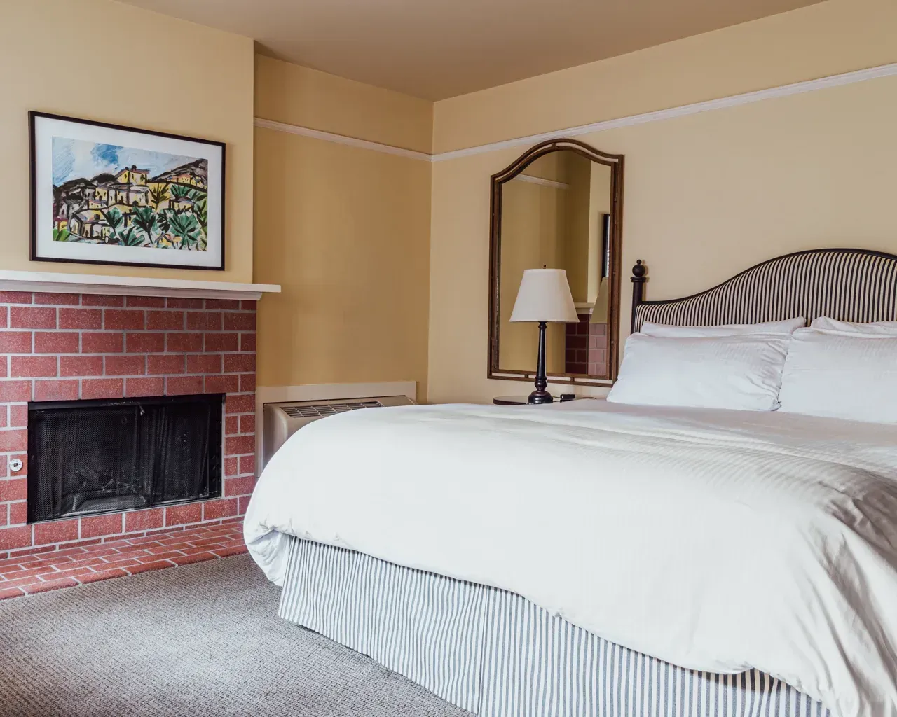 A hotel bedroom with a bed featuring striped bedding, a brick fireplace, and a framed painting on the wall.