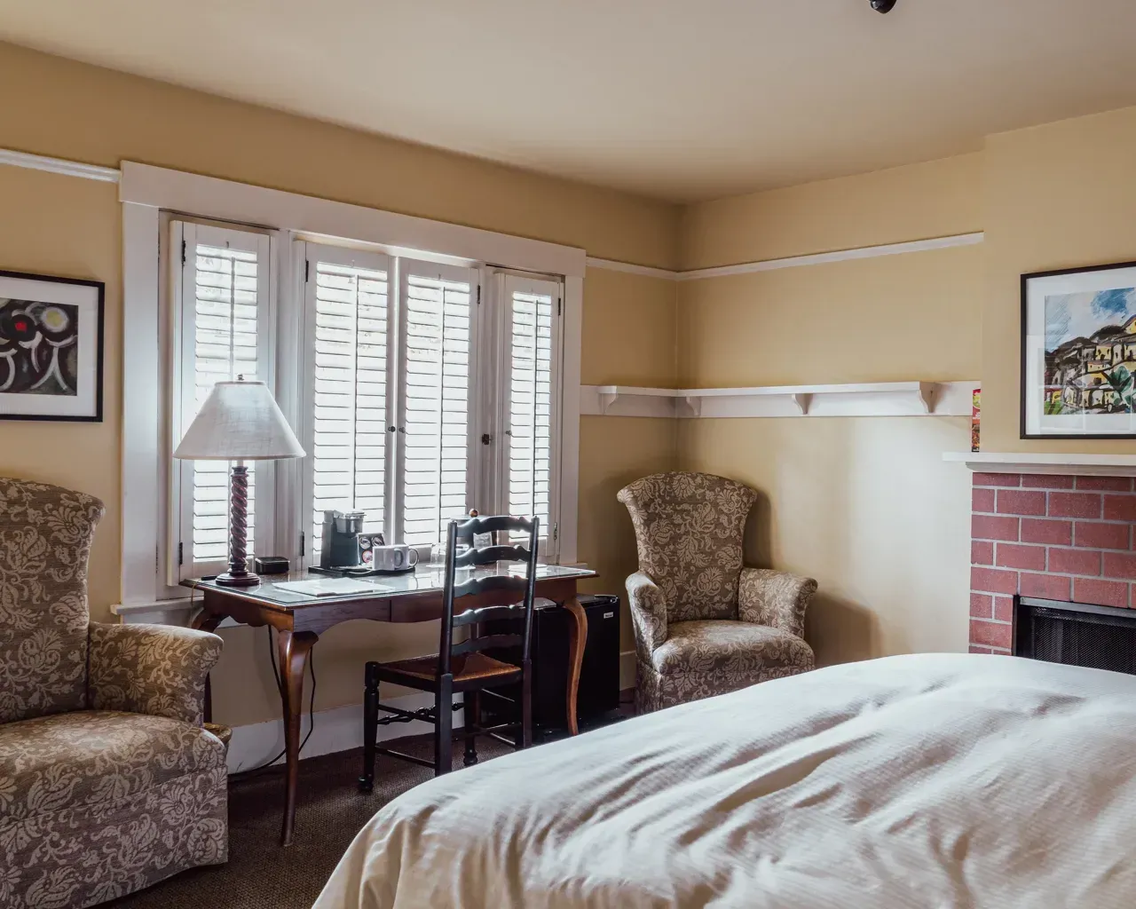 A bedroom with yellow walls, a bed, two patterned armchairs, and a wooden desk by a window with white shutters.