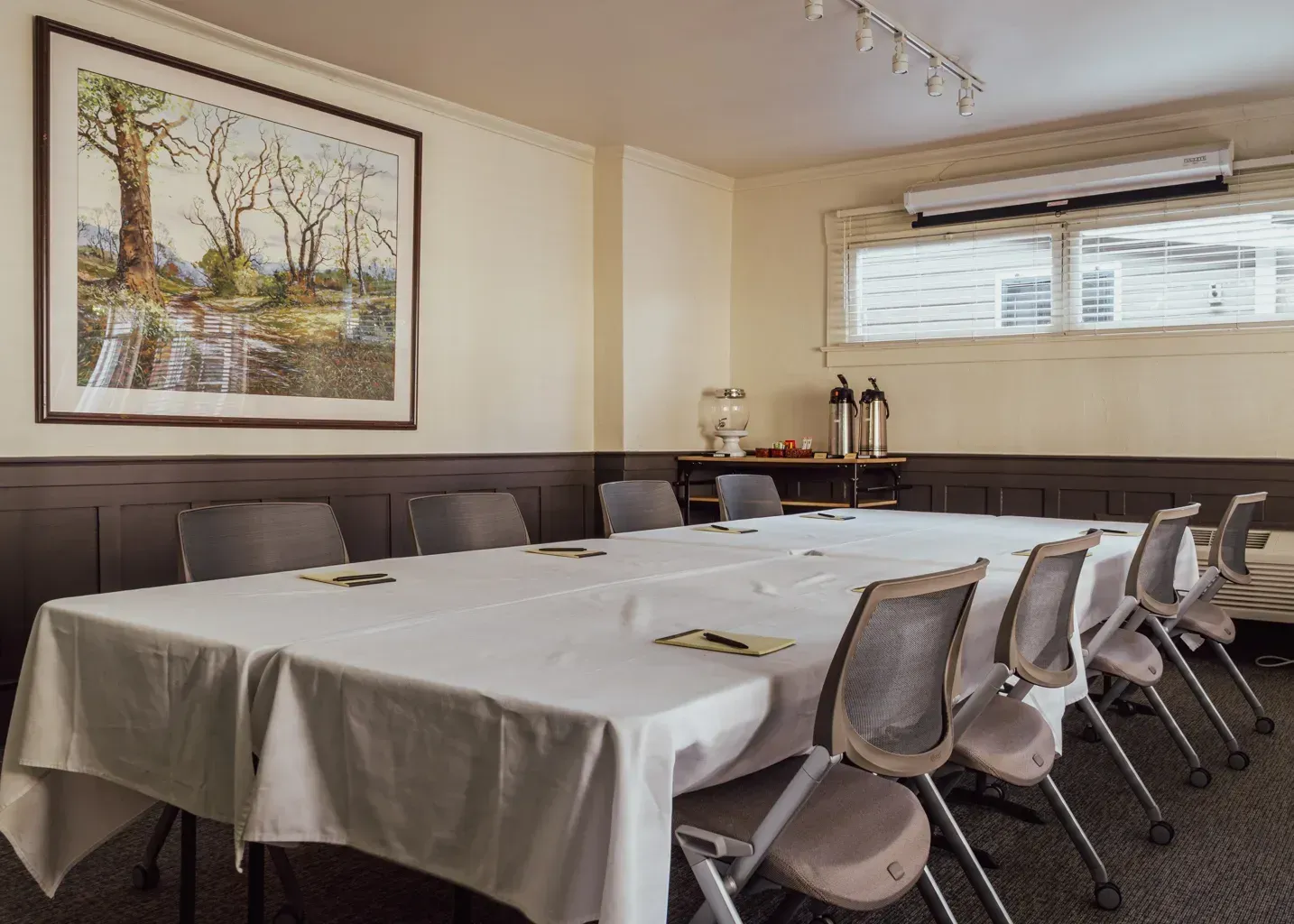 A long table with a white tablecloth and chairs set up in a conference room with a large framed landscape painting.