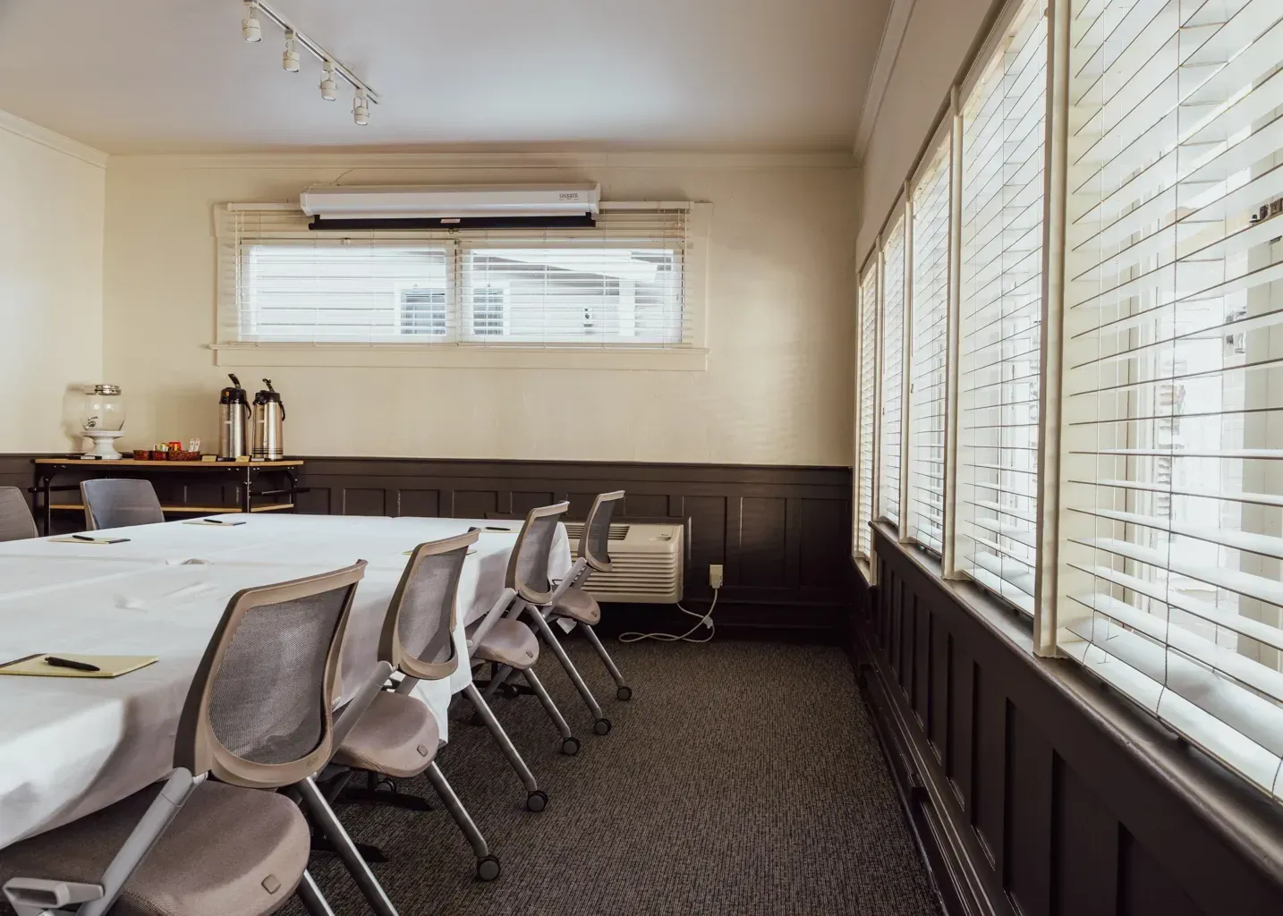 A conference room with a long table, chairs, and large windows with horizontal blinds along the side wall.