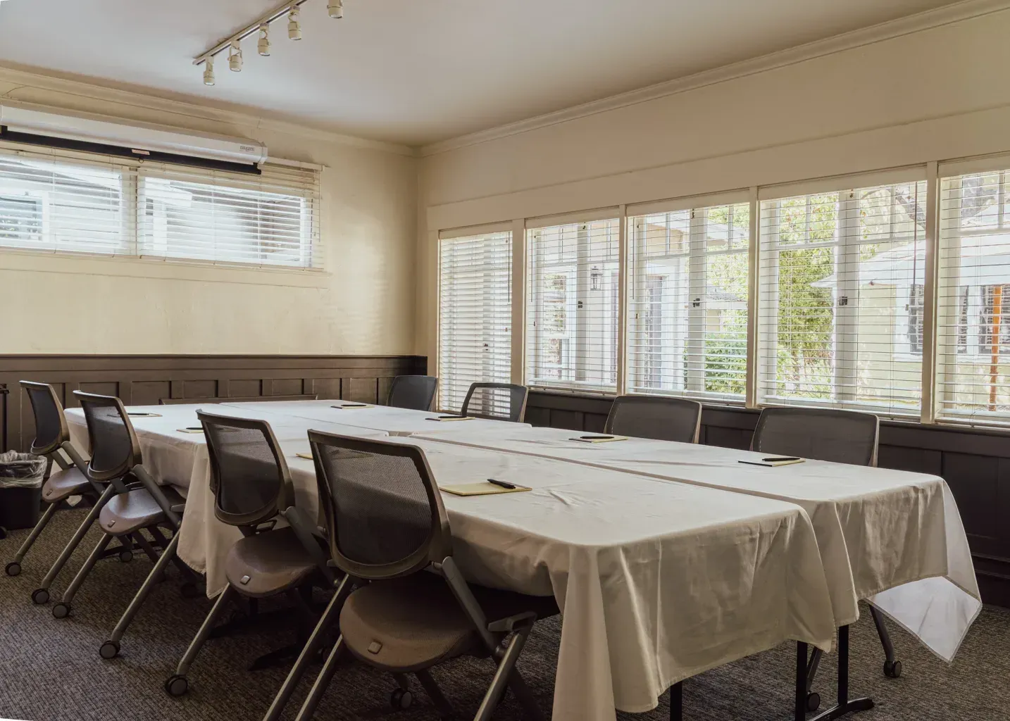 A conference room with two long tables draped in white cloths, surrounded by several grey office chairs.