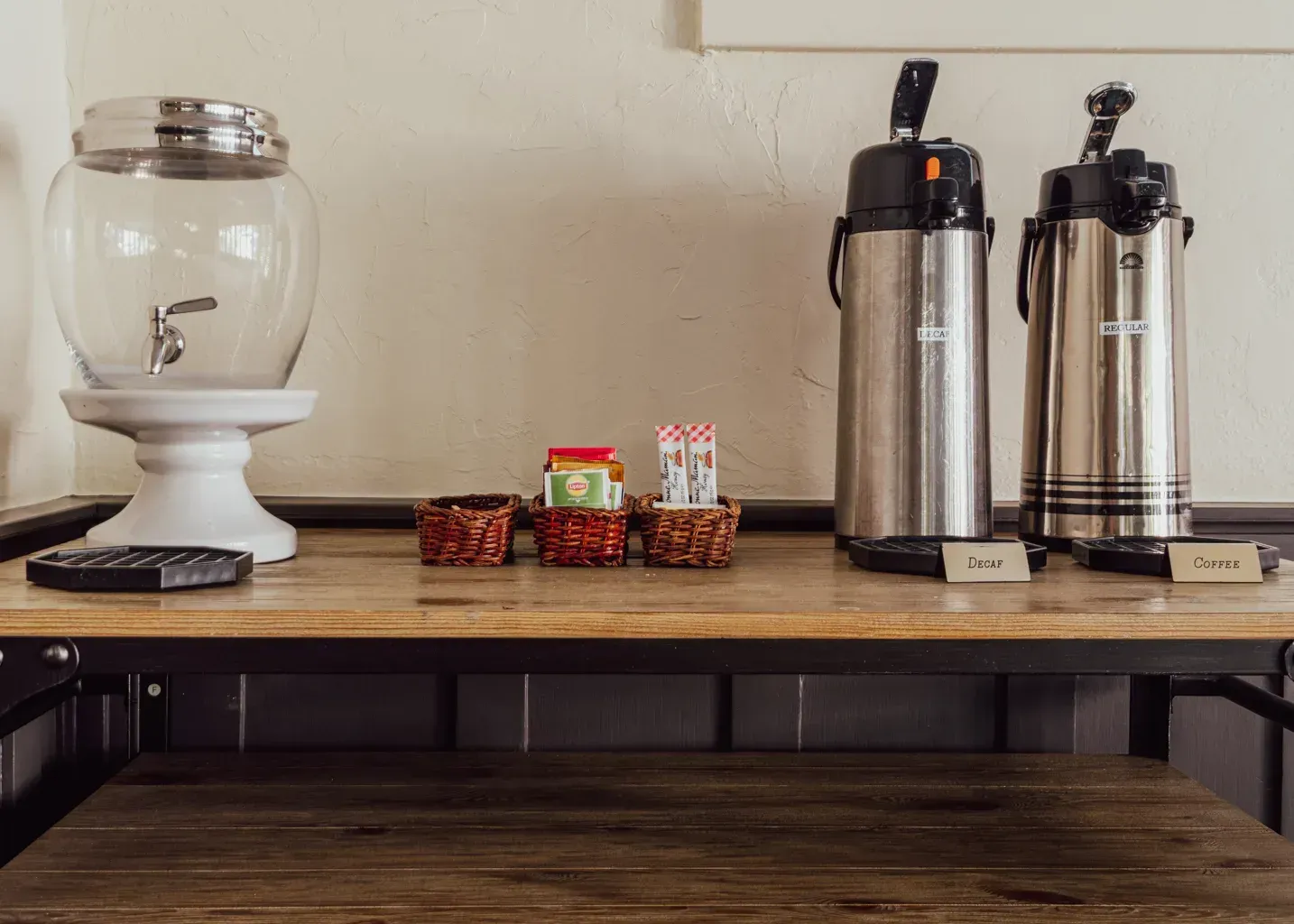 A beverage station with a glass water dispenser, coffee carafes, and small containers of tea bags on a wooden table.