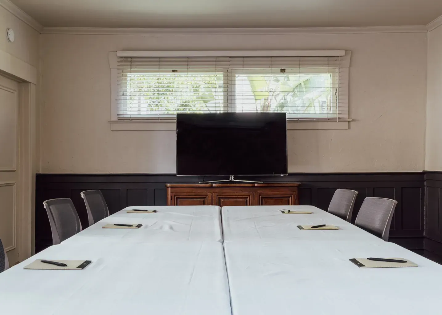 A meeting room with a long table covered by white cloths, surrounded by gray chairs, facing a television on a cabinet.