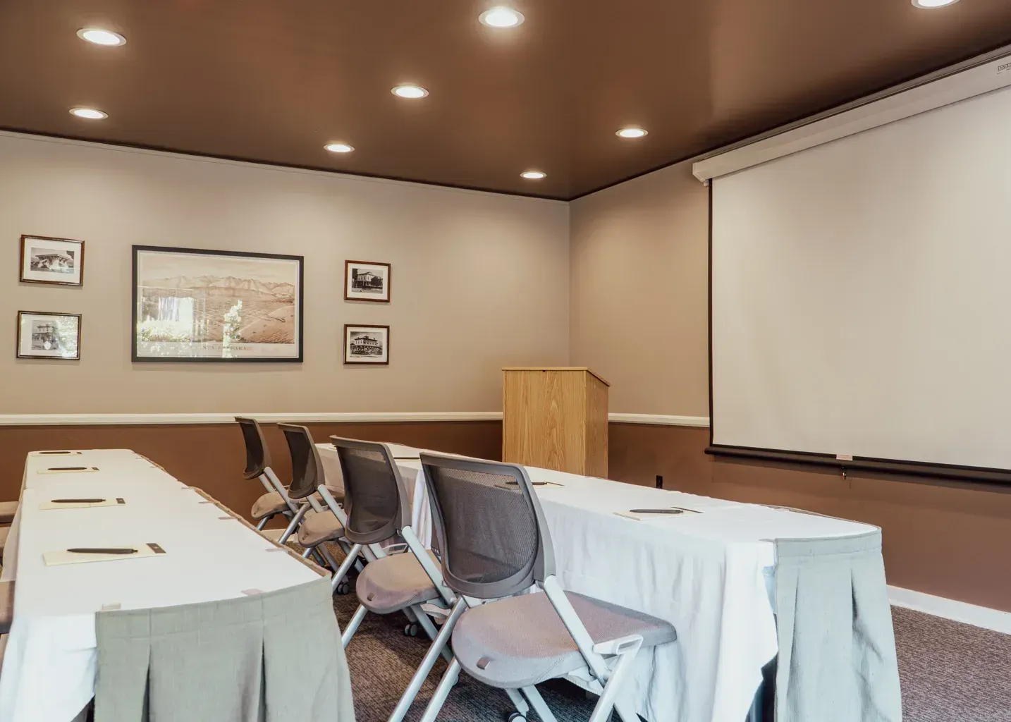 A conference room with two rows of tables and chairs, a wooden podium, and a large blank projector screen.