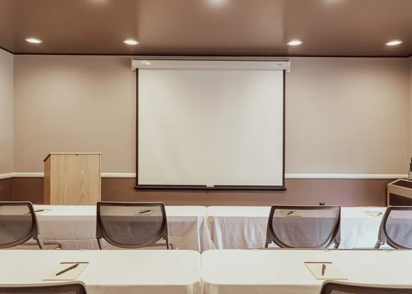 A beige meeting room with rows of tables, grey chairs, and a central projection screen behind a wooden lectern.