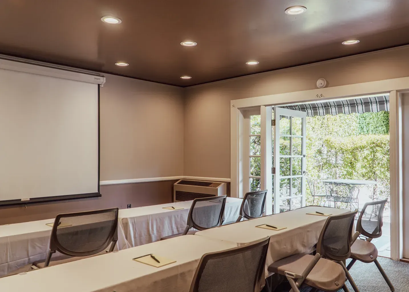 A conference room with rows of tables and chairs facing a projection screen, with open doors leading to a green patio.