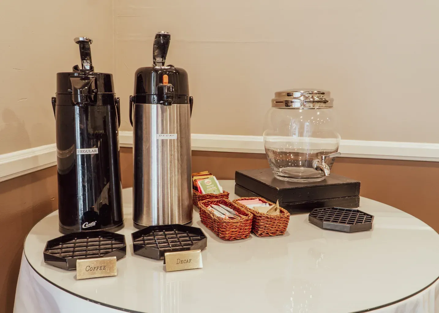 A coffee station with two thermal carafes, a glass container, and small baskets of supplies on a white round table.