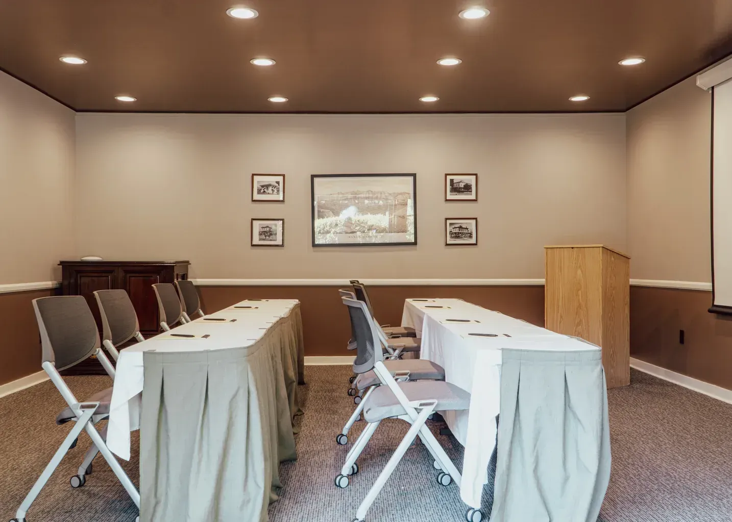 A conference room set with two rows of tables draped in white cloths, grey chairs, and a wooden podium against the wall.