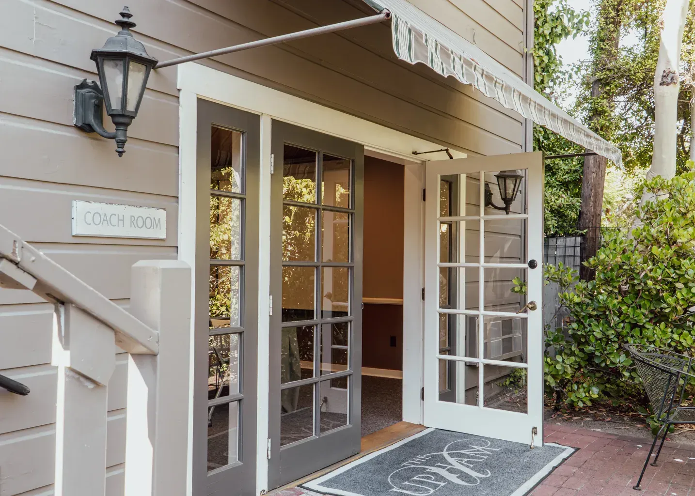 A brown building exterior with an open glass-paneled door, a wall-mounted lantern, and a welcome mat on a brick patio.