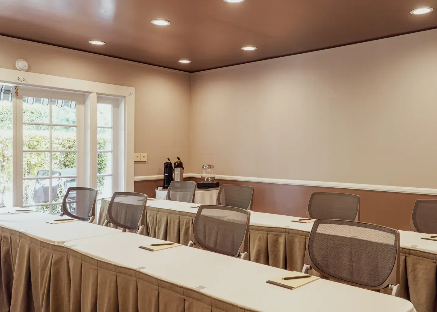 A meeting room with long tables covered in beige tablecloths, rows of gray office chairs, and recessed ceiling lights.