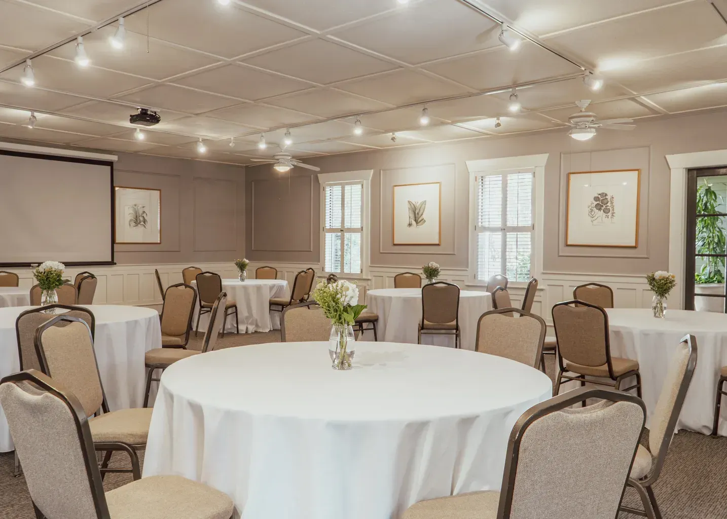 A well-lit meeting room featuring several round tables with white tablecloths and chairs, arranged for an event.