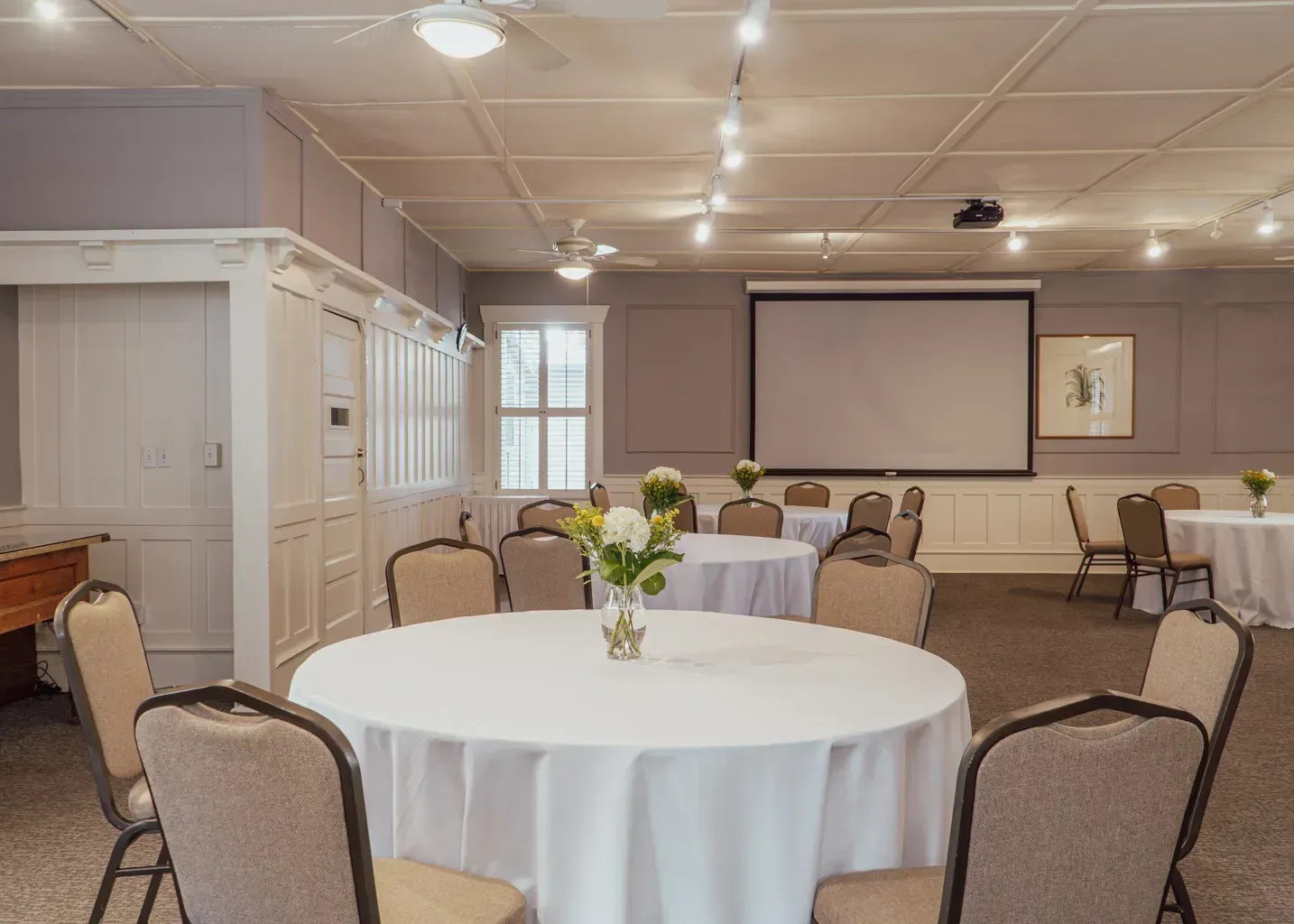 A bright event room with several round tables covered in white tablecloths, chairs, and a projection screen.