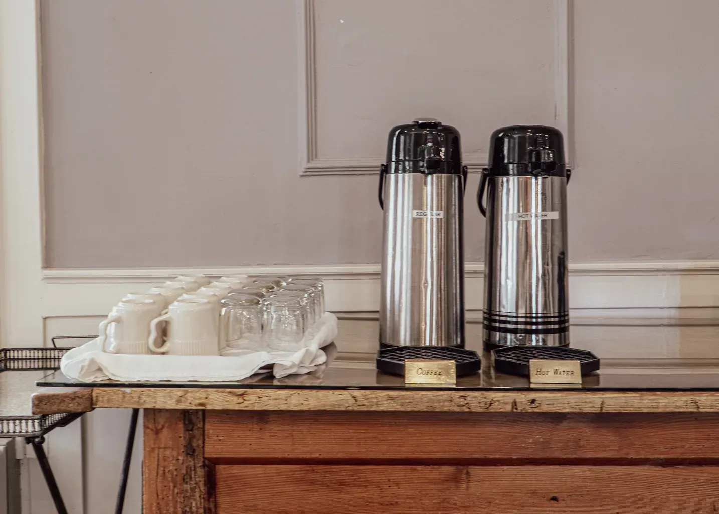 Two stainless steel coffee thermoses and a tray of small white mugs on a rustic wooden table against a paneled wall.