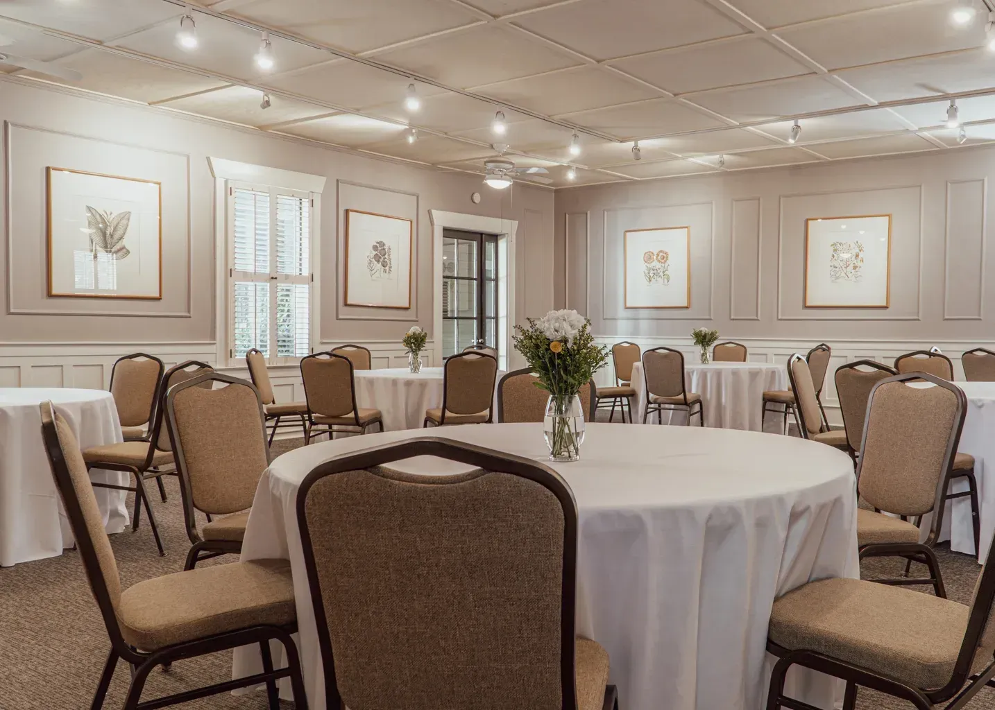 A formal event space with round tables covered in white tablecloths, beige chairs, and framed botanical art on the walls.