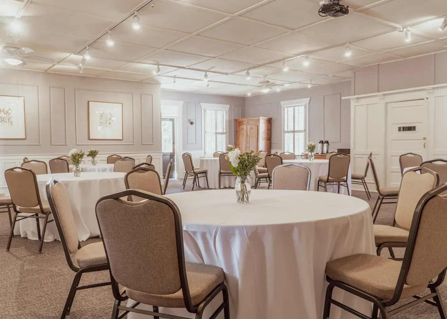 Event hall with round tables and chairs, light beige walls, neutral decor, and ceiling track lighting.