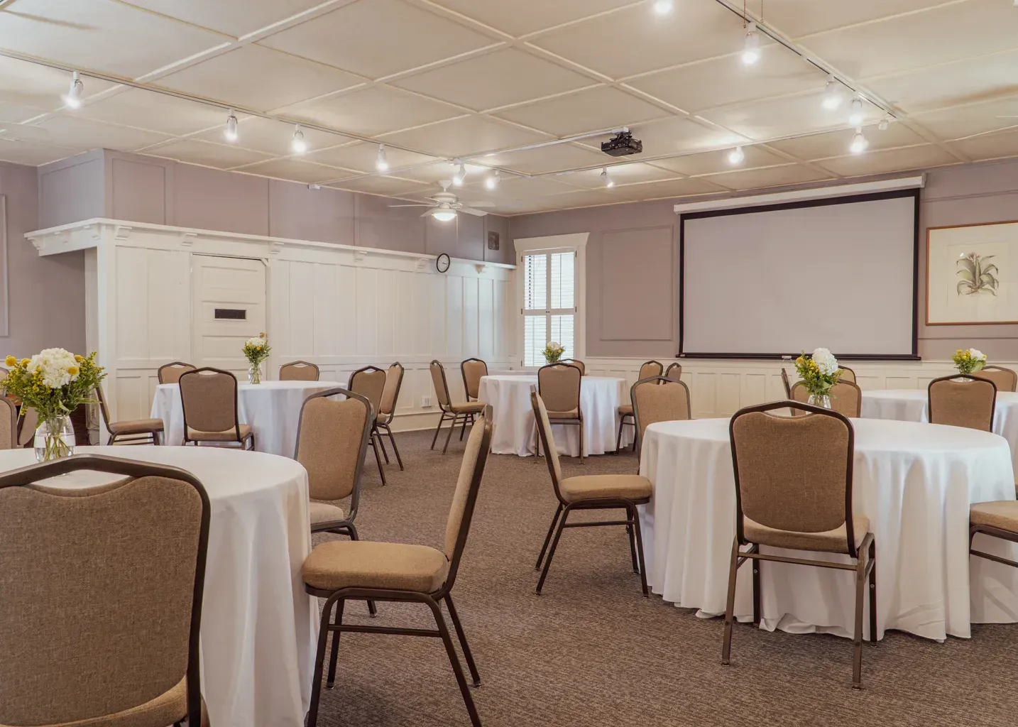 A bright, neutral-toned conference room with round tables, chairs, and a large projector screen.