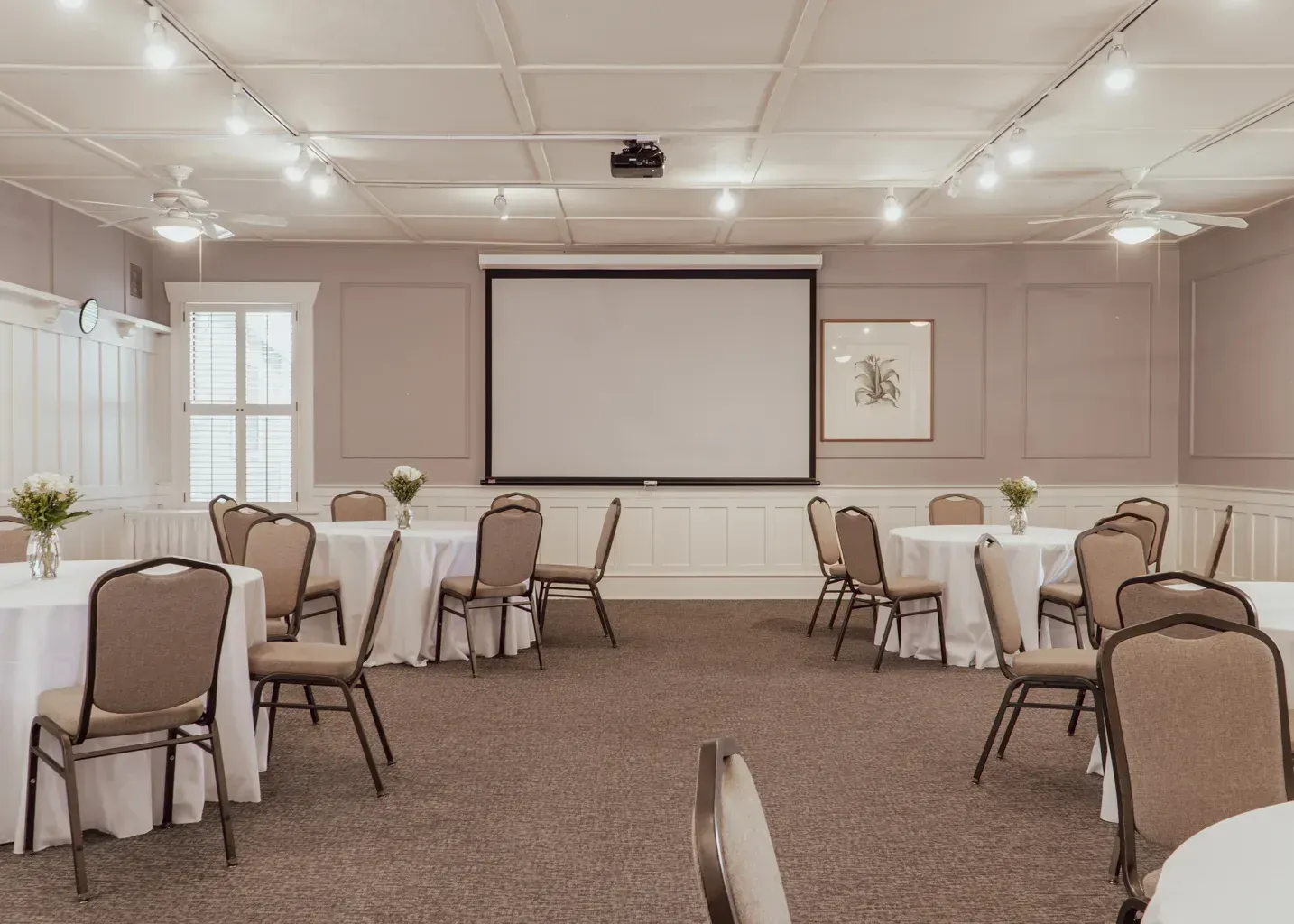 A meeting room with round tables, chairs, and a large projector screen centered on a light-colored wall.