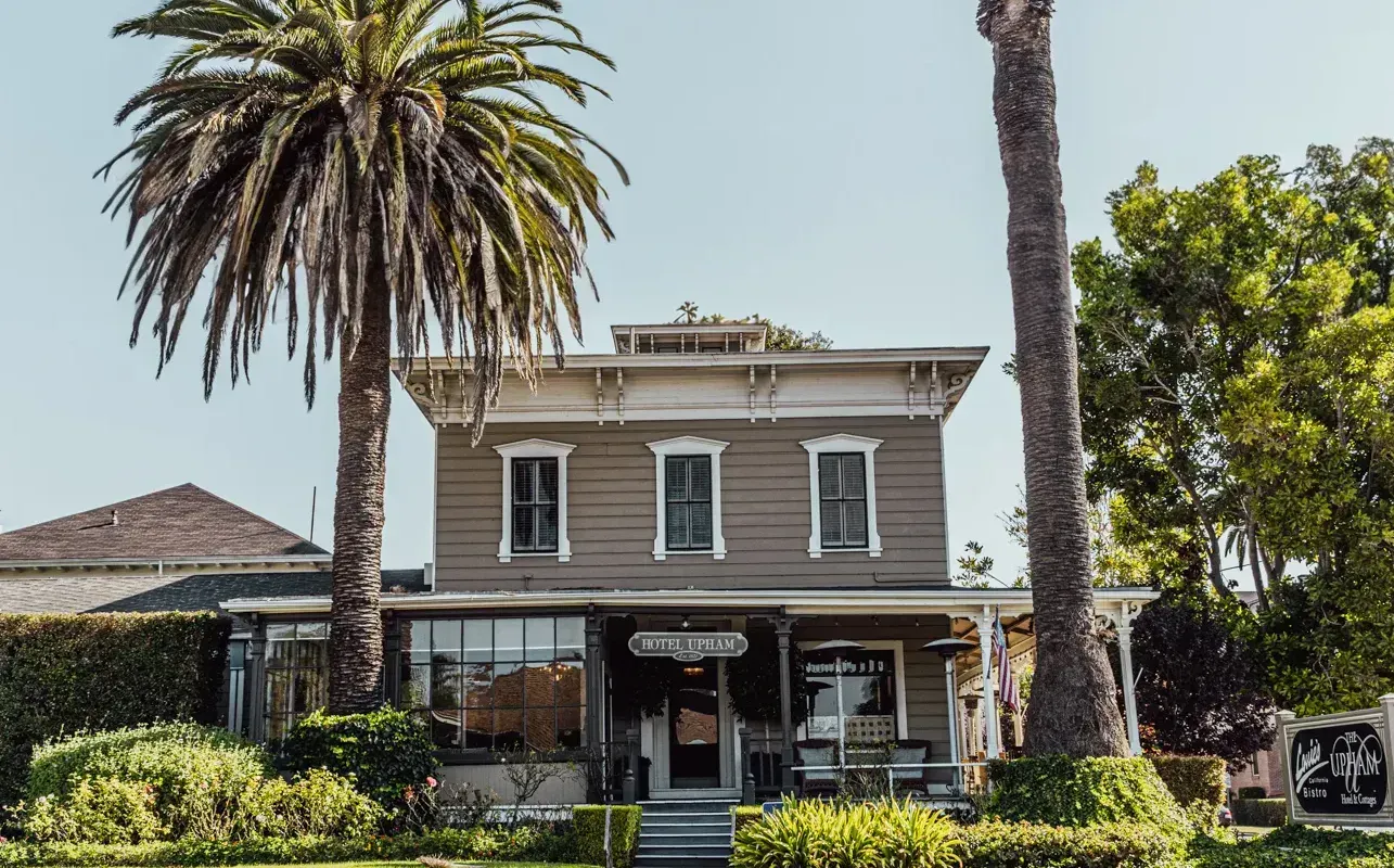 A two-story, tan-colored wooden historic building with a front porch, flanked by tall palm trees under a clear sky.