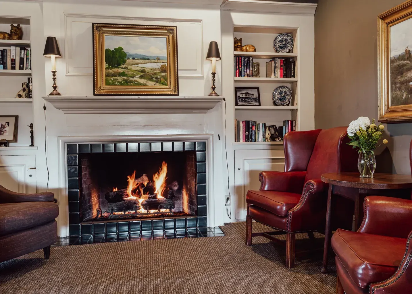 A cozy room with a lit fireplace, flanked by white bookshelves and two red leather wingback chairs.