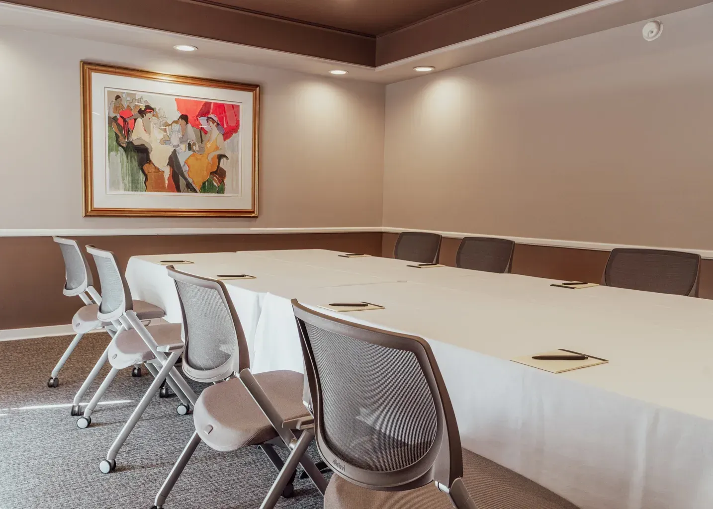 A conference room with a long table covered in a white cloth, surrounded by modern gray chairs, with art on the wall.
