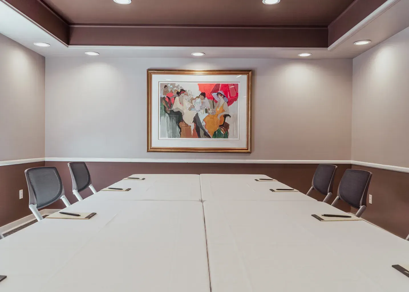 A modern conference room with a long table covered in a white cloth and black chairs, featuring a framed abstract print.