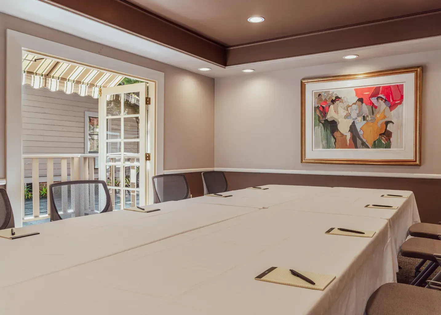 A long conference table with notebooks and pens set for a meeting in a room with a painting and door leading to a patio.