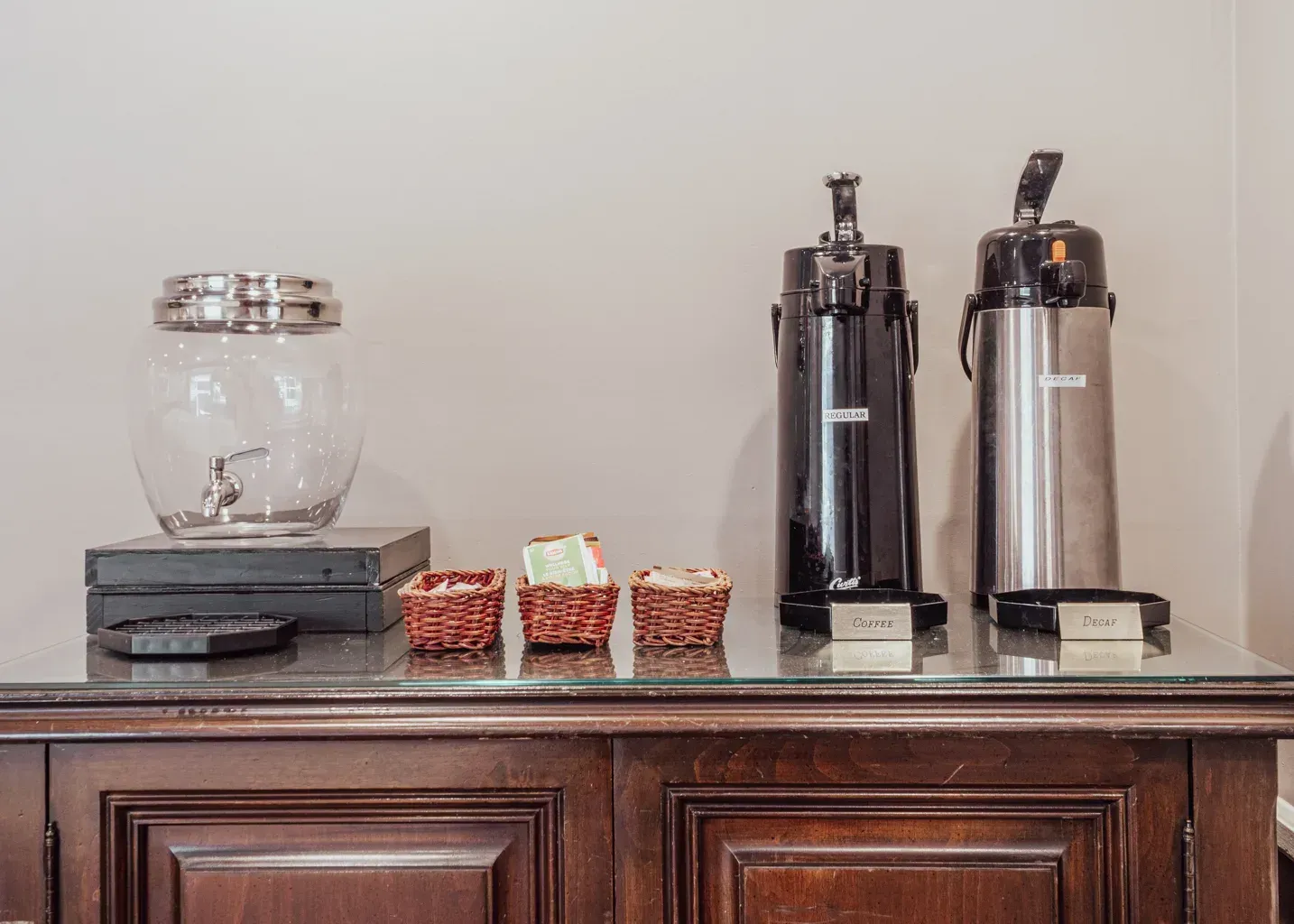 A self-service refreshment station with a glass water dispenser, three small baskets of items, and two thermal carafes.