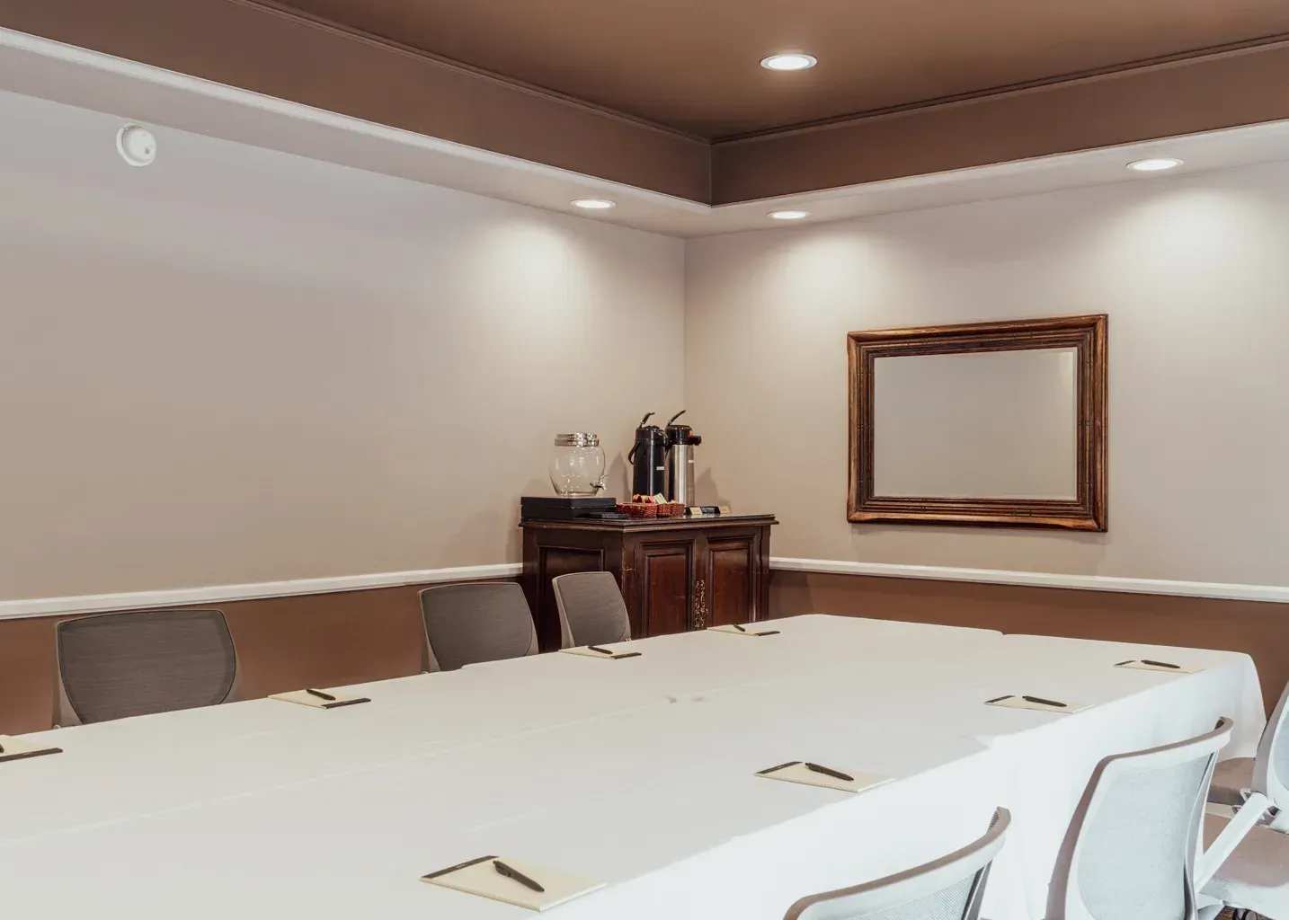 A small, modern meeting room with a long table covered in a white tablecloth, grey chairs, and a beverage station.