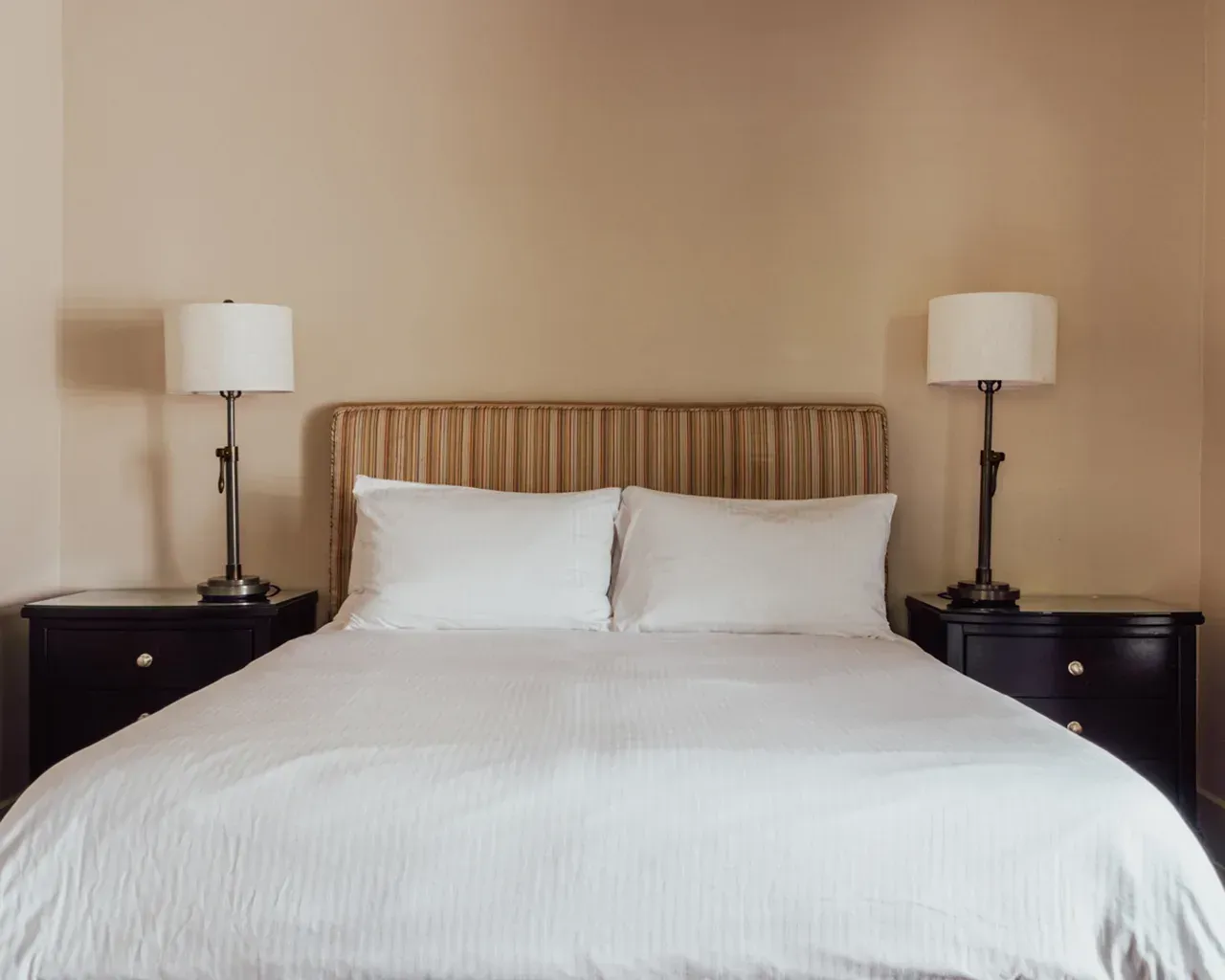 A neatly made bed with white bedding, a tufted beige headboard, and matching lamps on dark brown nightstands.