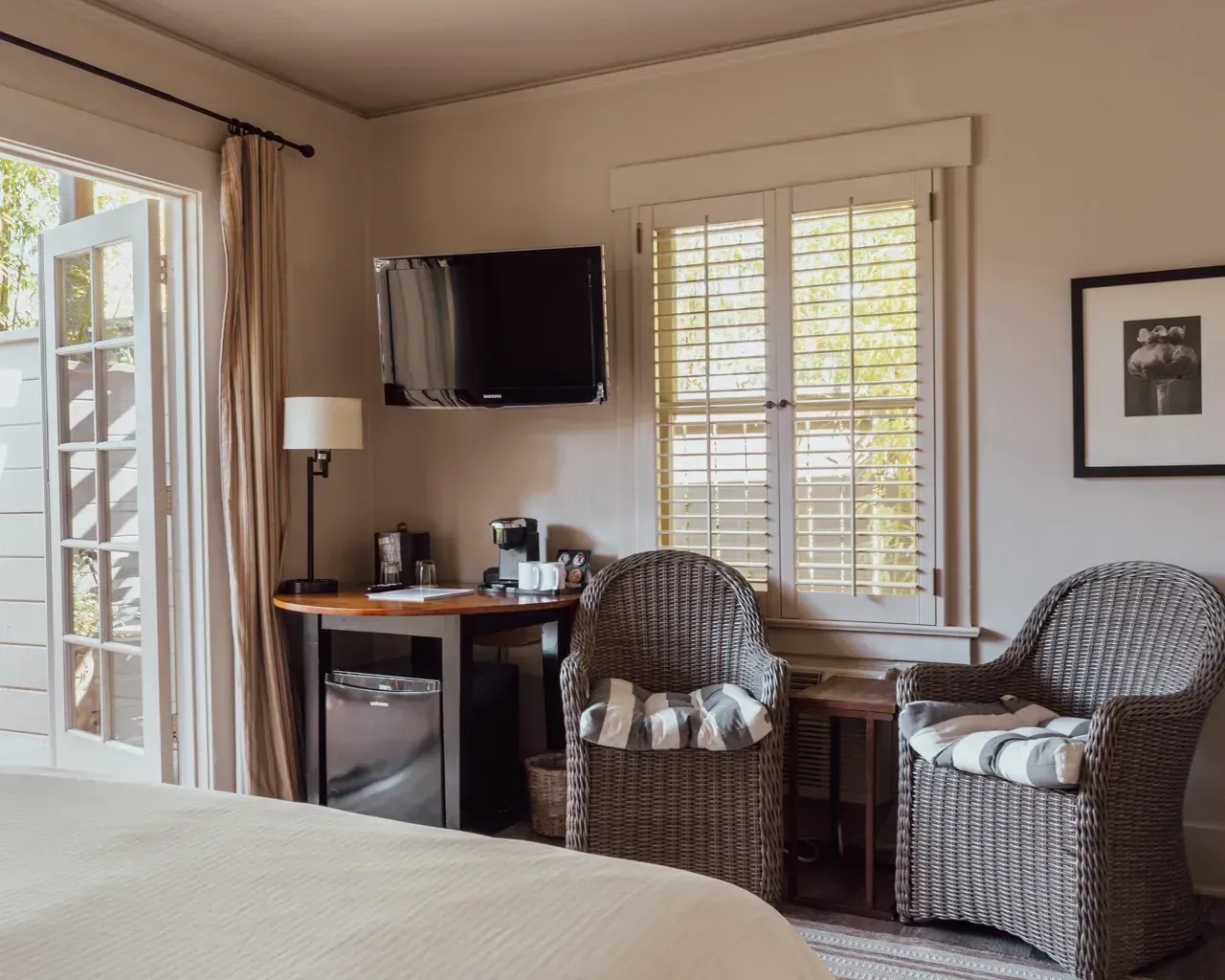 A hotel room featuring two wicker chairs, a small table with a coffee machine, and a wall-mounted television.