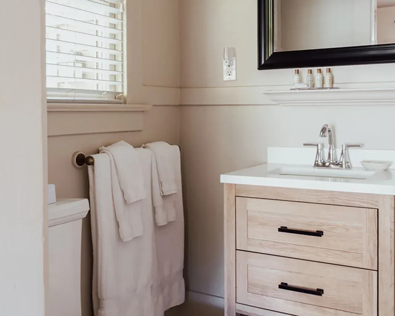 A bathroom vanity with a light wood cabinet, white countertop, sink, mirror, and white towels hanging on the wall.