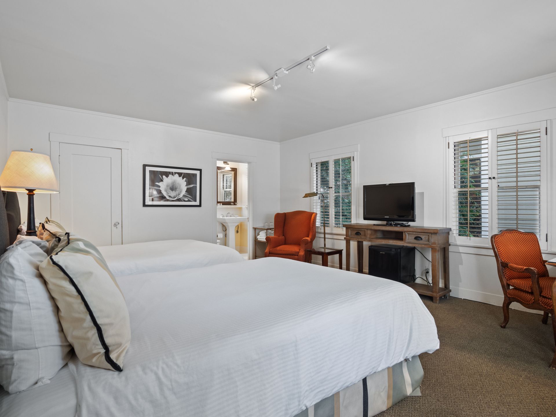 A hotel bedroom with two beds, a lamp, a television on a wooden stand, two orange chairs, and a window with blinds.
