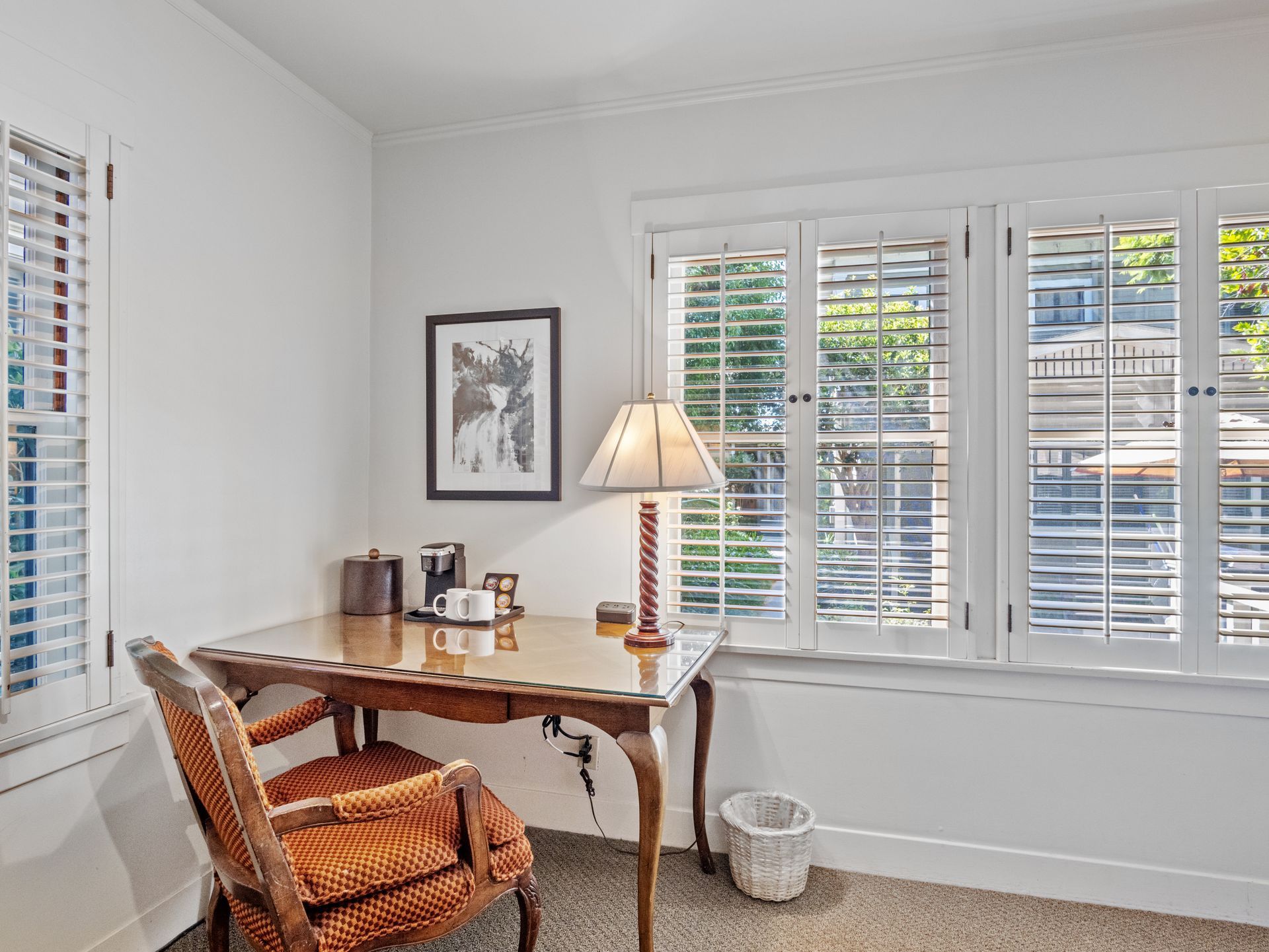 A wooden desk with a lamp, chair, and coffee maker sits in a bright room with white walls and shuttered windows.