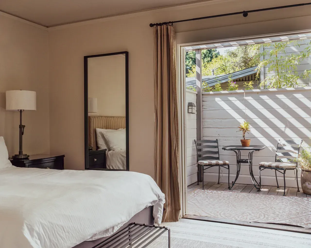 A bedroom with a bed and lamp next to a glass sliding door opening to a private patio with a small table and two chairs.