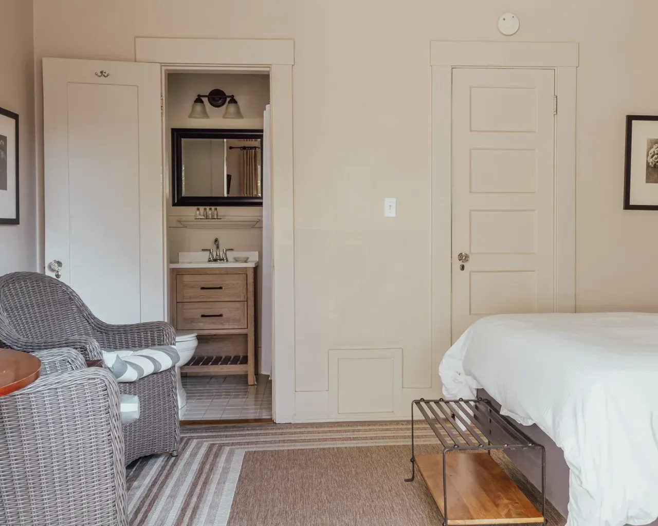 A bedroom with a wicker armchair, a bed with white bedding, and an open doorway revealing a small bathroom vanity.