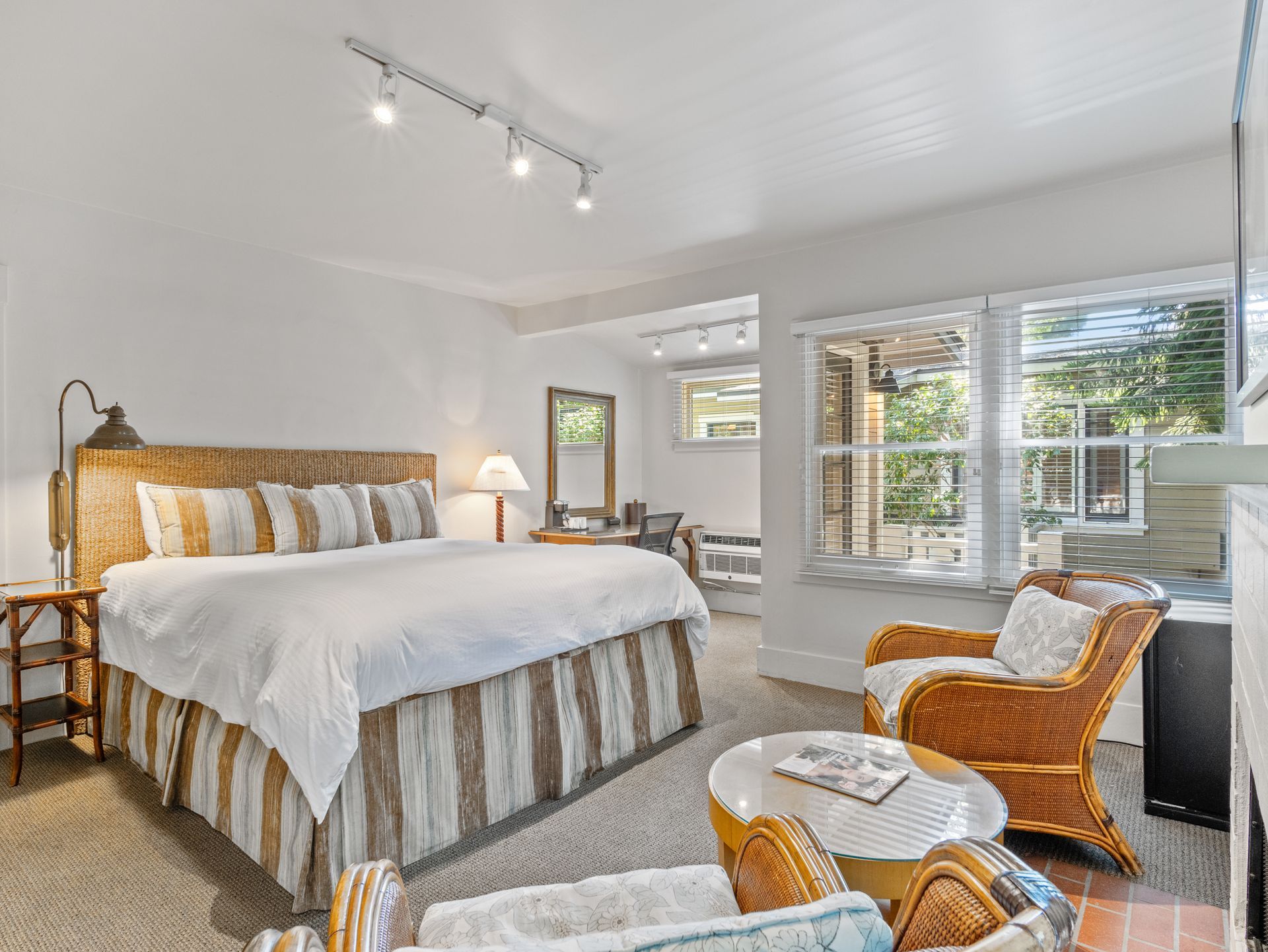 A hotel bedroom with a large bed, two wicker armchairs, a small coffee table, and a desk area by the window.