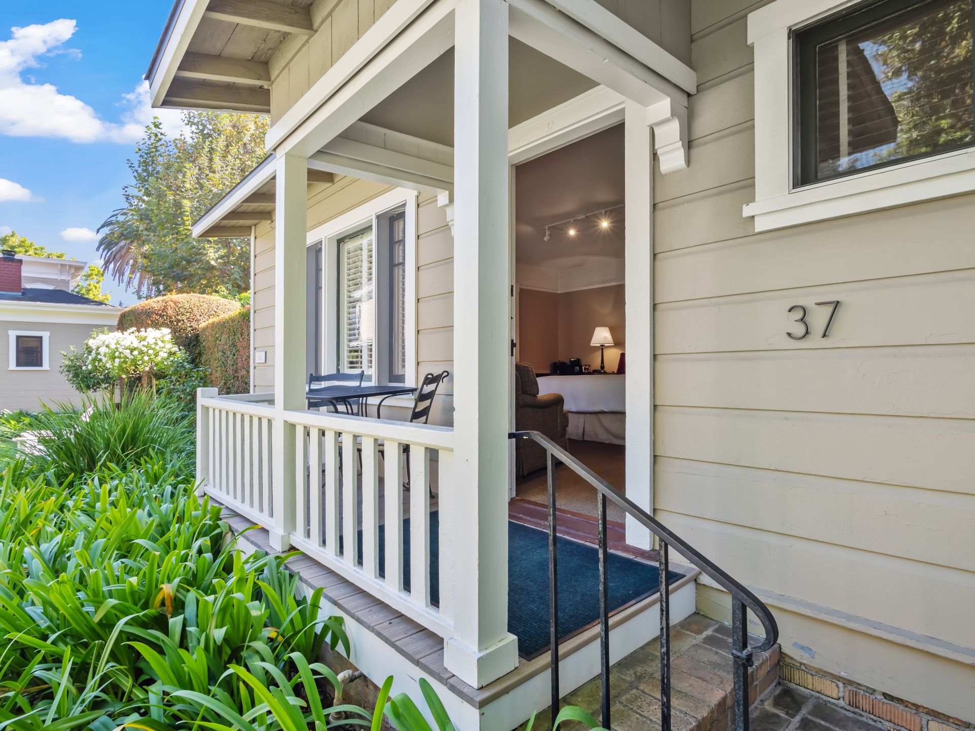 A tan house with a porch and white railings, featuring the number 37 near the front door and lush green garden foliage.