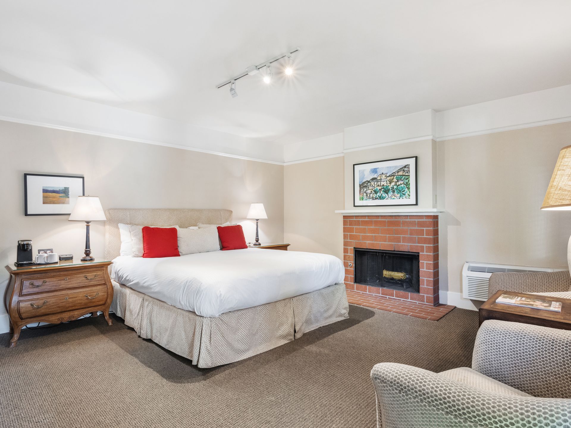 A hotel bedroom with a king bed, red accent pillows, a brick fireplace, a wooden dresser, and a gray armchair.